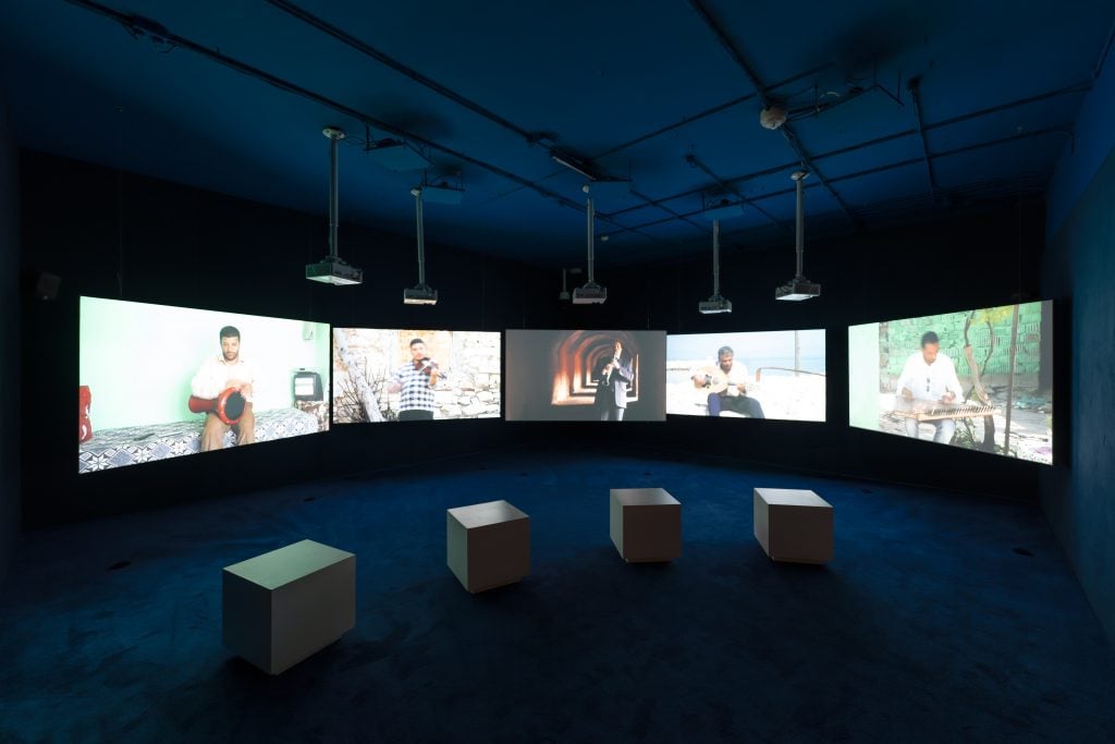 Five screen video installation by Ergin Çavuşoğlu in "Seeing is Believing" in Doha, exploring the influence of Gérôme and Orientalism. A five screen video installation wherein each has one musician playing music, which has then been synced together. Four square stools are in the center.