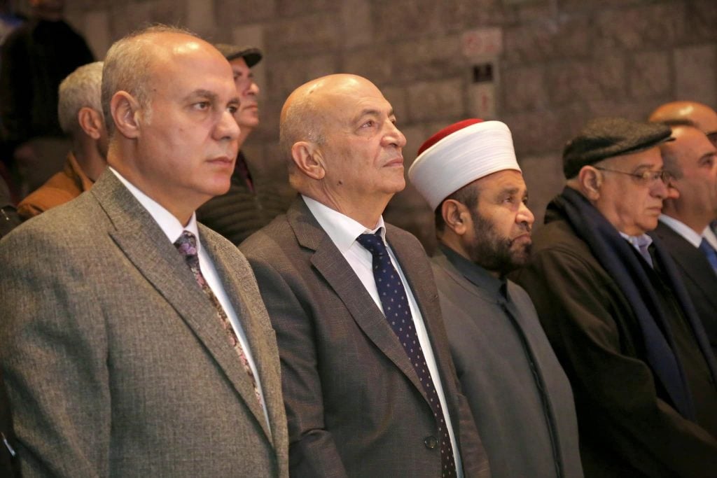 A group of men in formal attire, including suits and ties, stand in a row, appearing to attend a solemn event. One individual is wearing a traditional white headdress with a red stripe, signifying religious or cultural significance. The setting appears to be indoors, with a stone wall in the background.