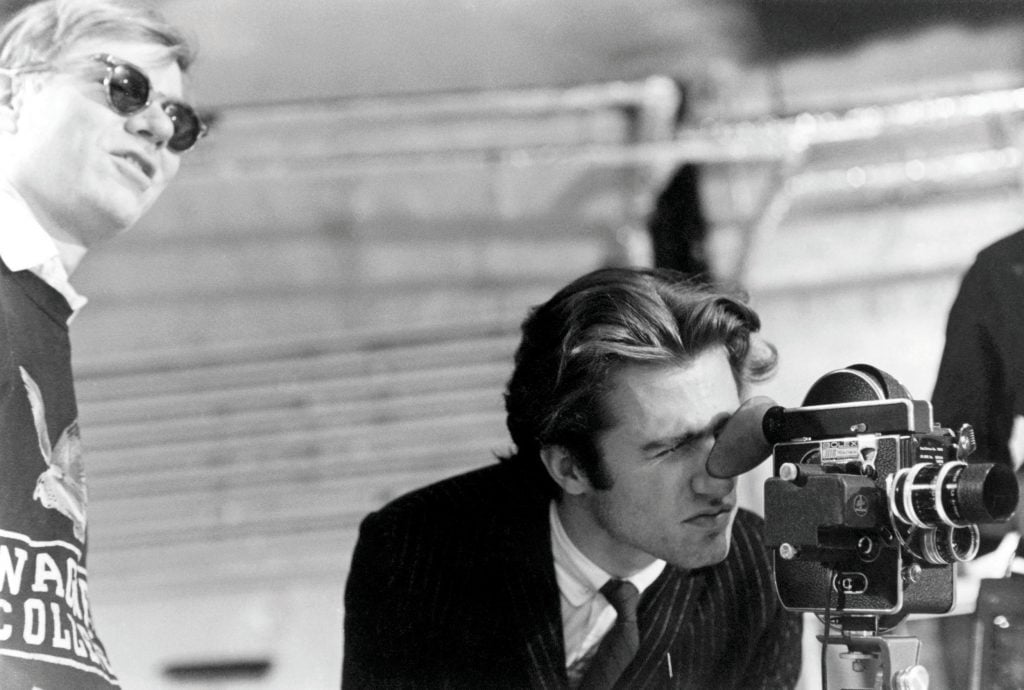 A man looking into a film camera, while Andy Warhol stands in the background