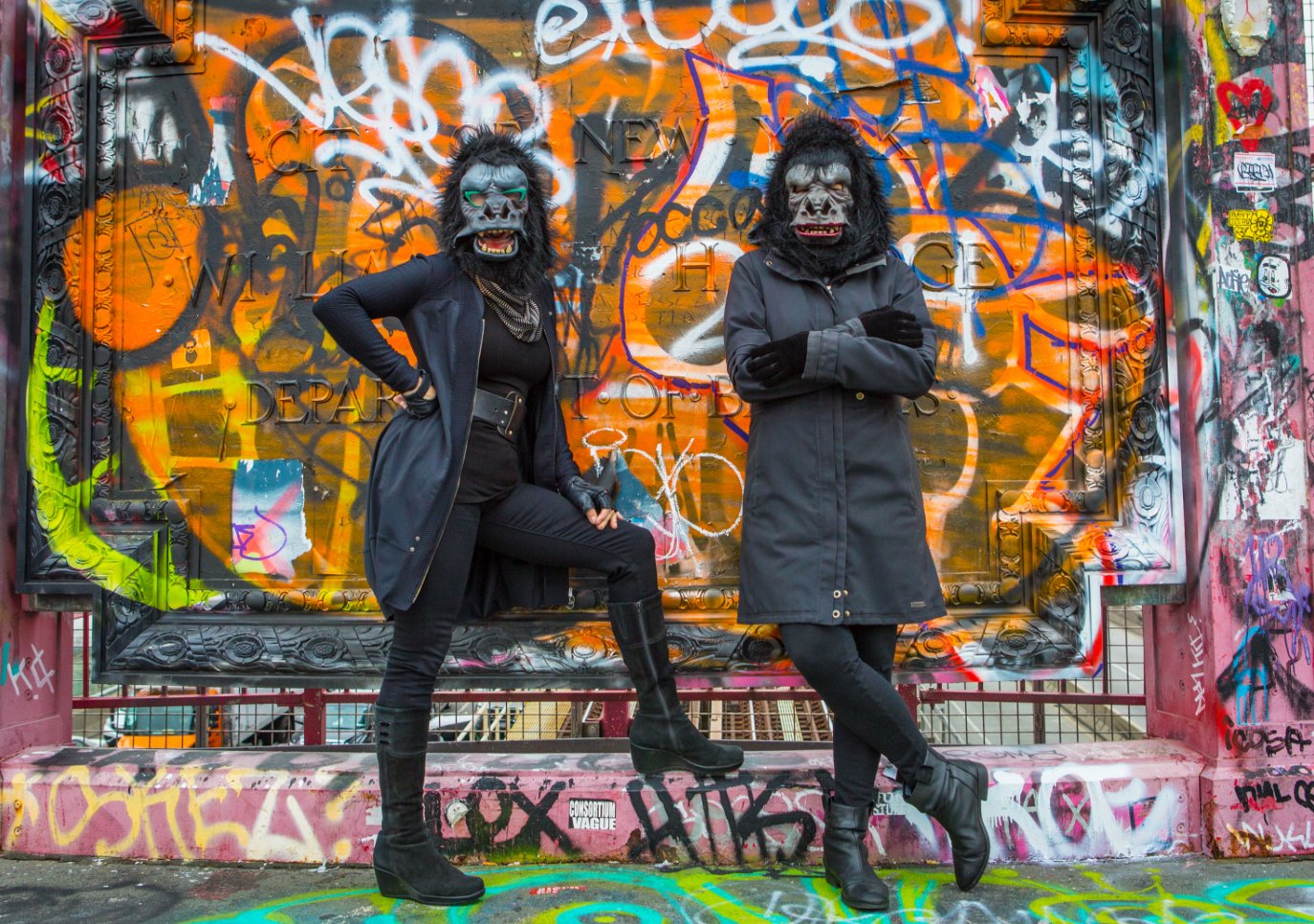 A photograph of two women dressed in all black and wearing gorilla masks standing before a heavily graffiti'ed wall.