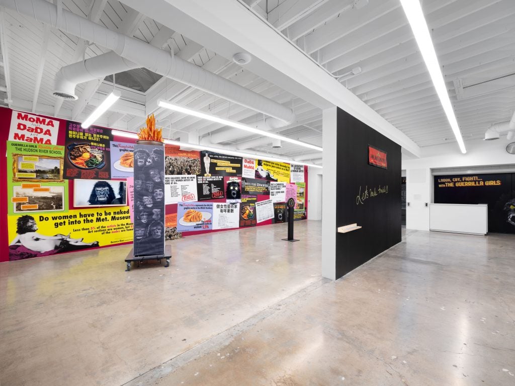 A photograph of the inside of a white walled gallery space, featuring one wall covered with colorful print outs, another in front of it covered in all black, and another free-standing artwork between them.