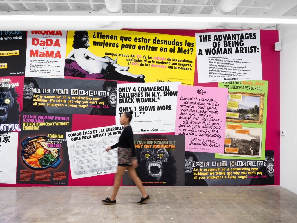 A photograph of a gallery wall covered with colorful digital print outs of art protest posters, with a woman walking in front of it.