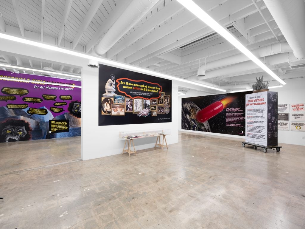 A photograph of a white walled gallery space featuring large-scale, printed posters and a small vitrine holding Guerilla Girls related ephemera at Beyond the Streets.