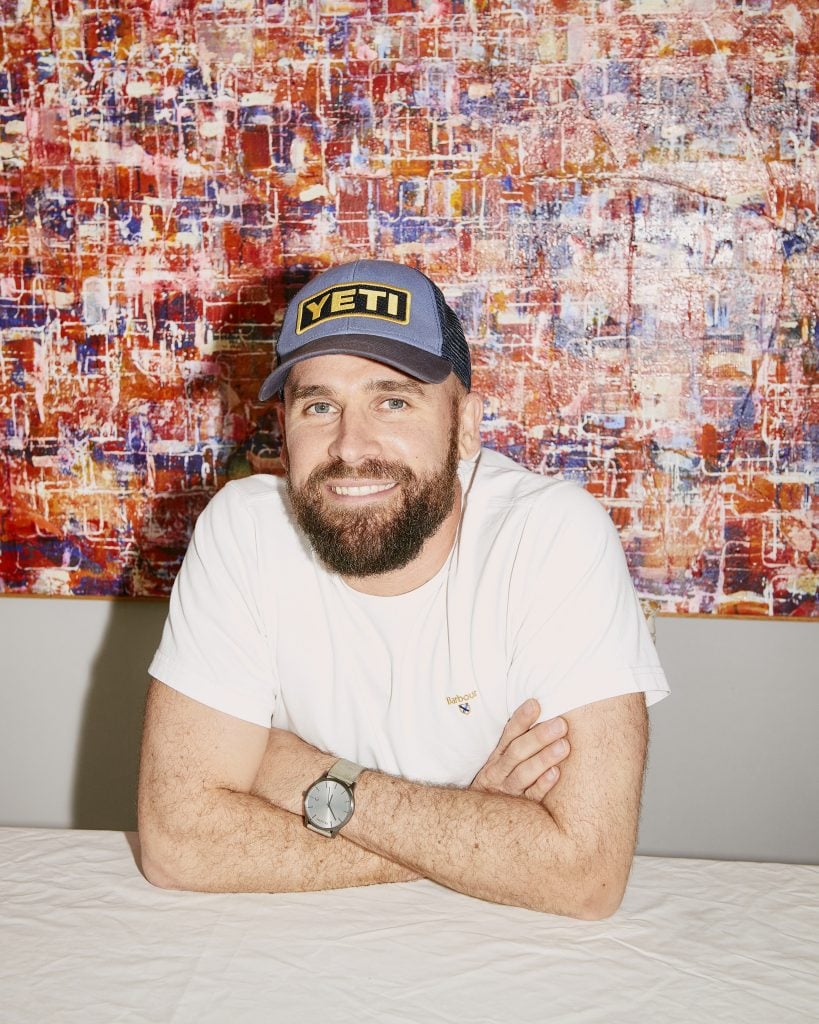 Portrait of Huw Lougher of Lougher Contemporary wearing a white t-shirt with his arms crossed at a white table, also wearing a Yeti baseball cap in front of an abstract artwork.