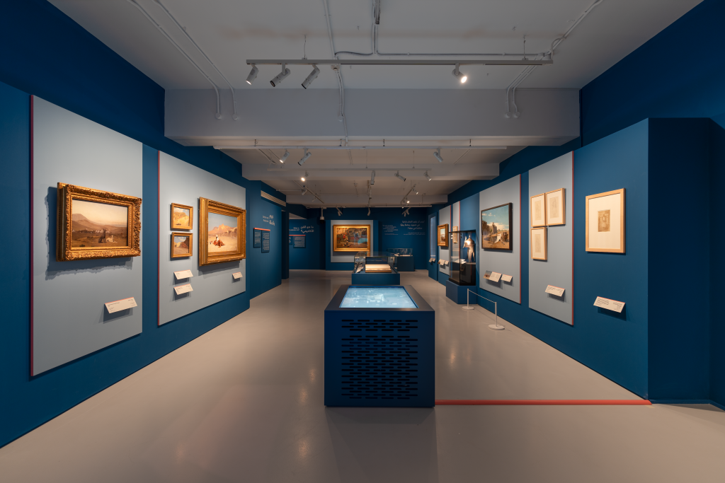 Installation view of Jean-Léon Gérôme solo exhibition at MATHAF in Doha, showing a long gallery space painted peacock blue with his paintings lining the walls and a video display flat in the center vitrine.