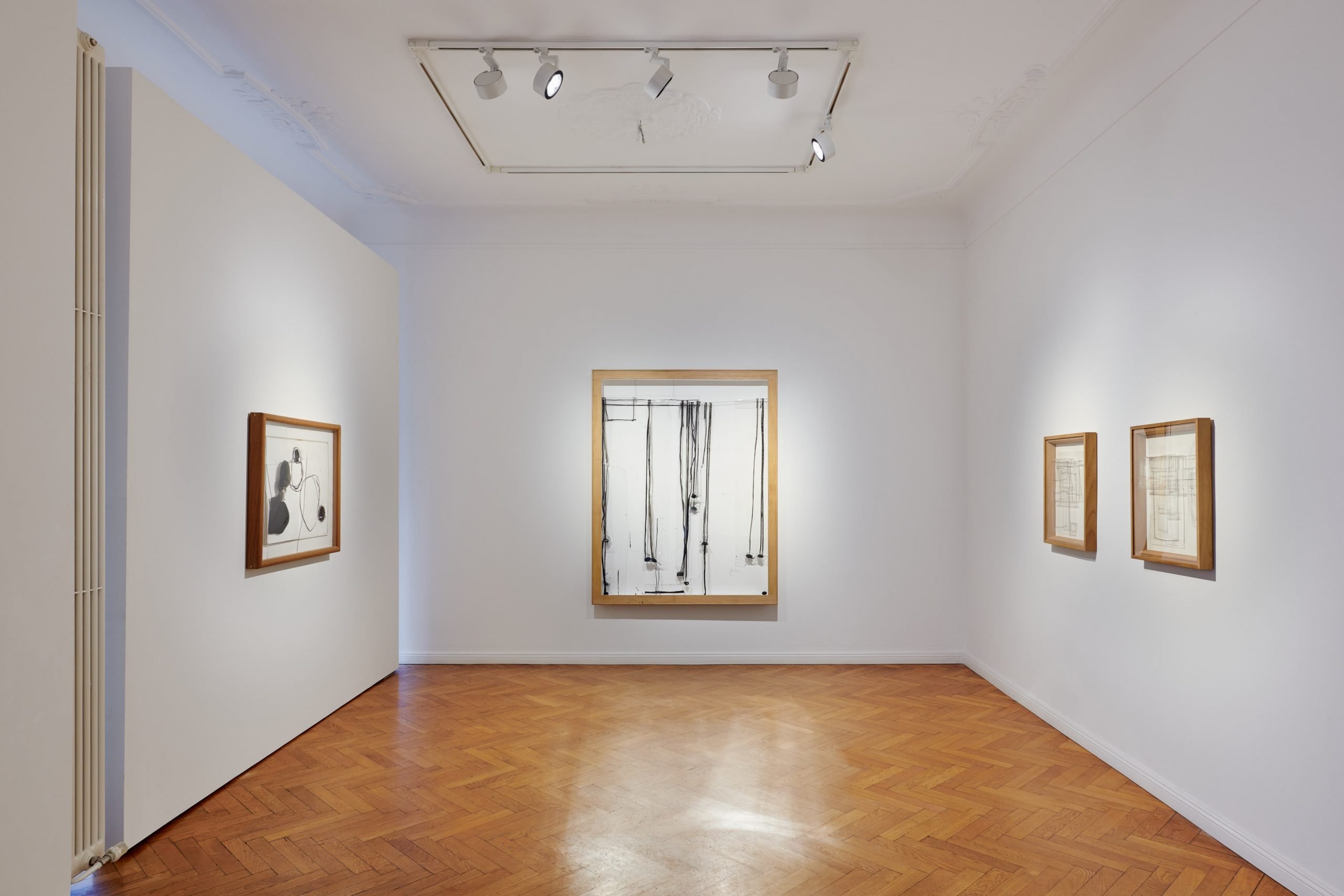 Installation view of the solo show "Jordi Alcaraz: Two Decades" showing a white gallery room space with a series of spotlighted works on paper and drawings framed on the white walls.