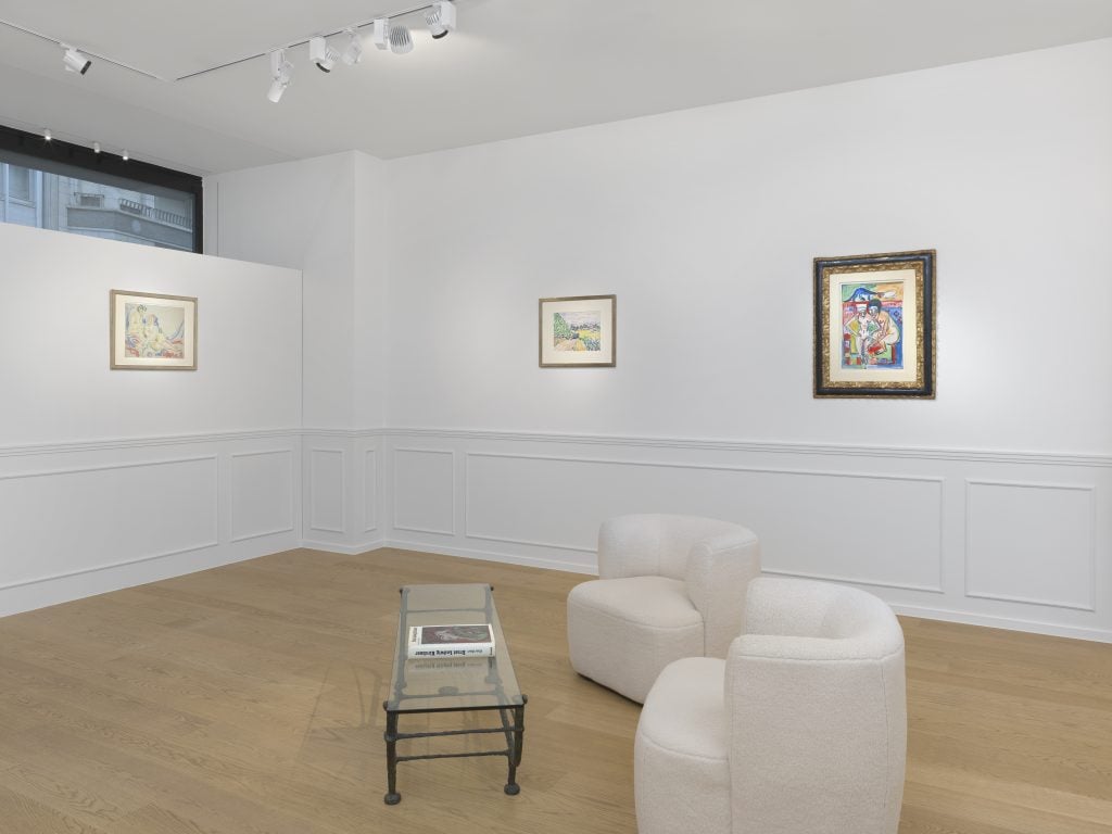 Installation view of Ernst Ludwig Kirchner exhibition at Bailly Gallery, showing the corner of a white gallery space with three artworks on the walls and a set of two upholstered chairs in the center and a small low table between them.