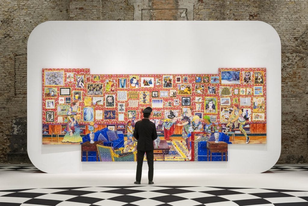 A person stands in front of a large-scale installation comprised of numerous, colorful small-scale flat works in a salon-wall style, standing on black and white checked tile floor.