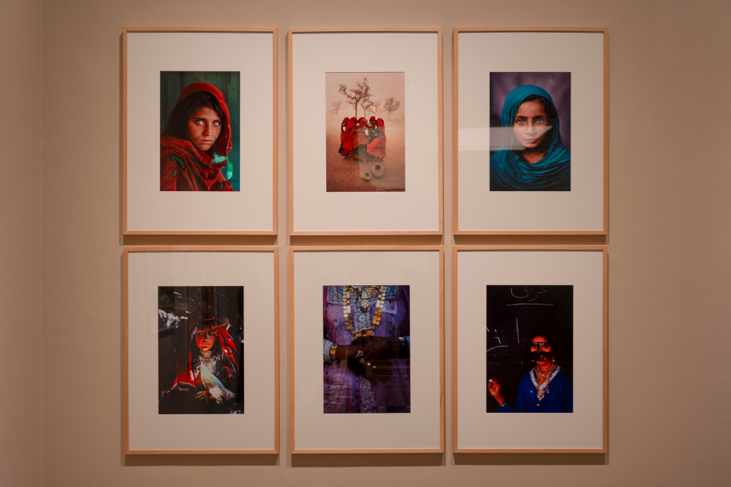 A suite of six framed photographs by Steve McCurry, four of which are portraits of MENASA women, one of which is a small group of people unseen behind red robes, and a closeup of a woman's clasped hands, all highlight saturated in color.