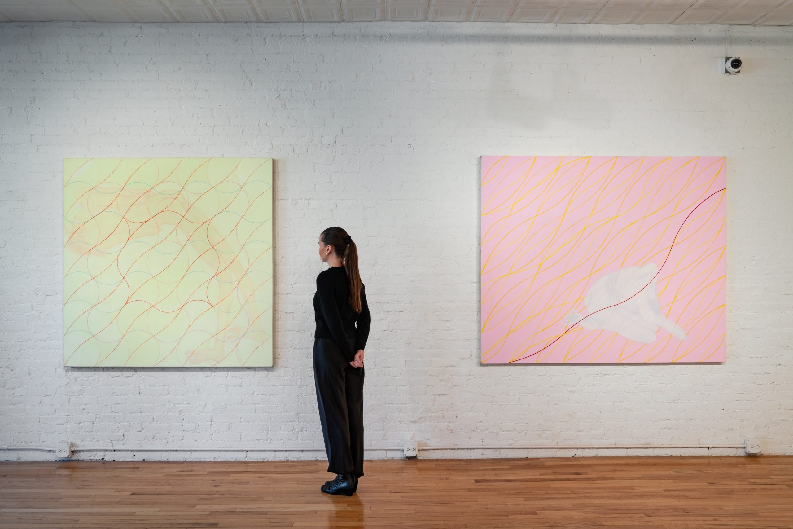 Installation view of "Audra Skuodas: Vibrational Vulnerability" (2024) at Cristin Tierney Gallery showing two pastel abstract paintings on a white wall with a woman in black standing in front of the one on the left looking at it.