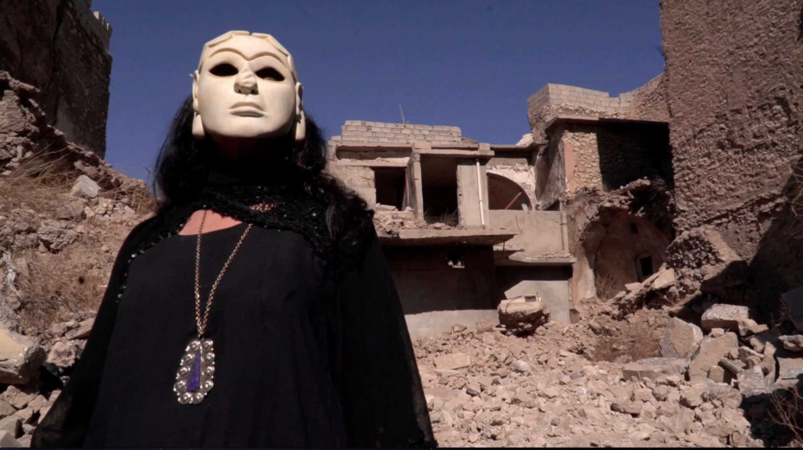 Film still from work by Ishtar Yasin Guriérrez whose work will be featured at Abu Dhabi Art 2024, showing a figure cloaked in black and wearing a white statue mask standing in front of what appears to be stone ruins.