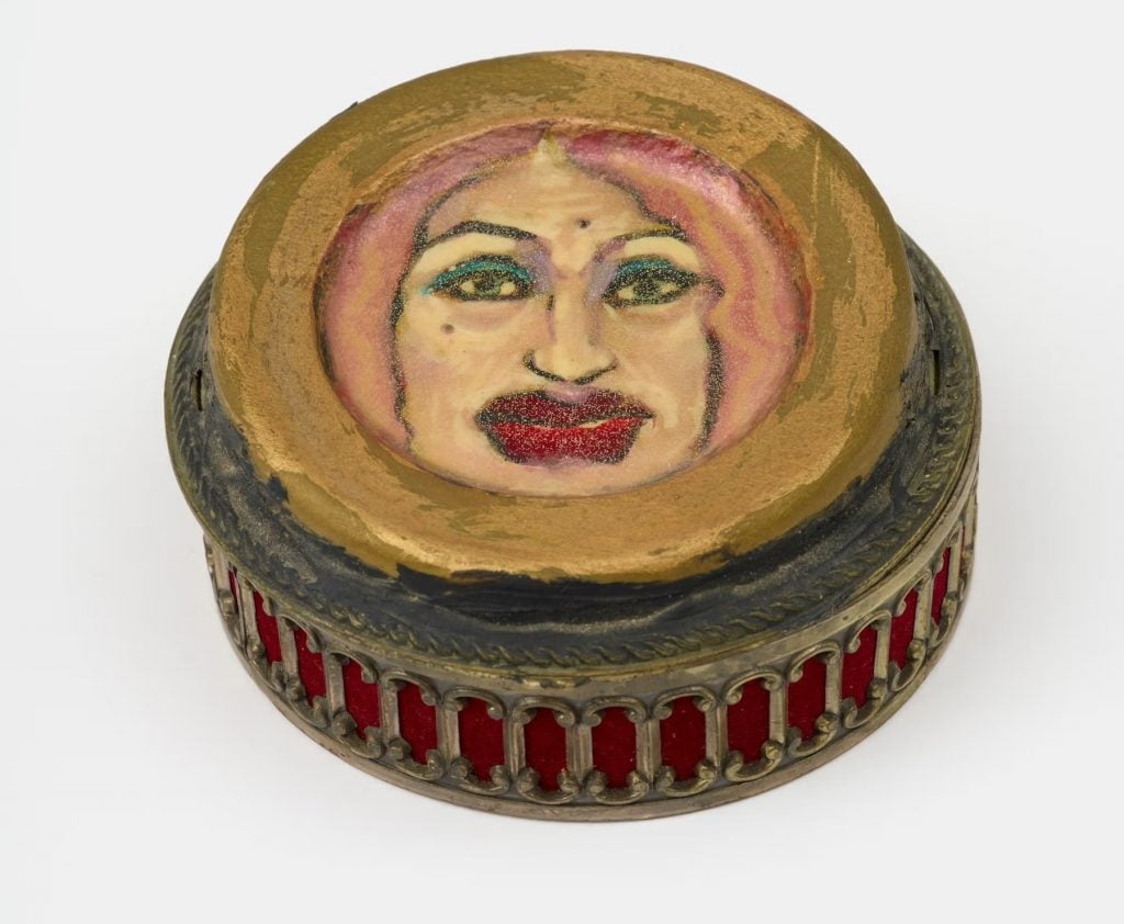 A close-up of a circular painting depicting a woman with pink hair, prominent eyebrows, and red lipstick. The painting is set within a gold frame and is mounted on a wooden base.