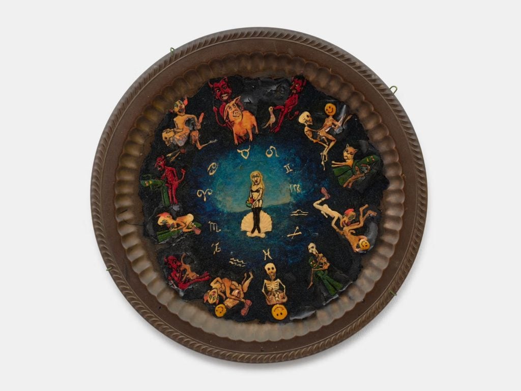 A circular metal plate with a dark brown border and a raised rim. The interior is painted with a dark blue background and features a central image of a figure with long blonde hair and a blue dress. The figure is holding a white object in her hands. Surrounding her are twelve smaller images depicting various zodiac signs, each with a unique figure or symbol. Some figures are human-like, while others are more abstract or symbolic