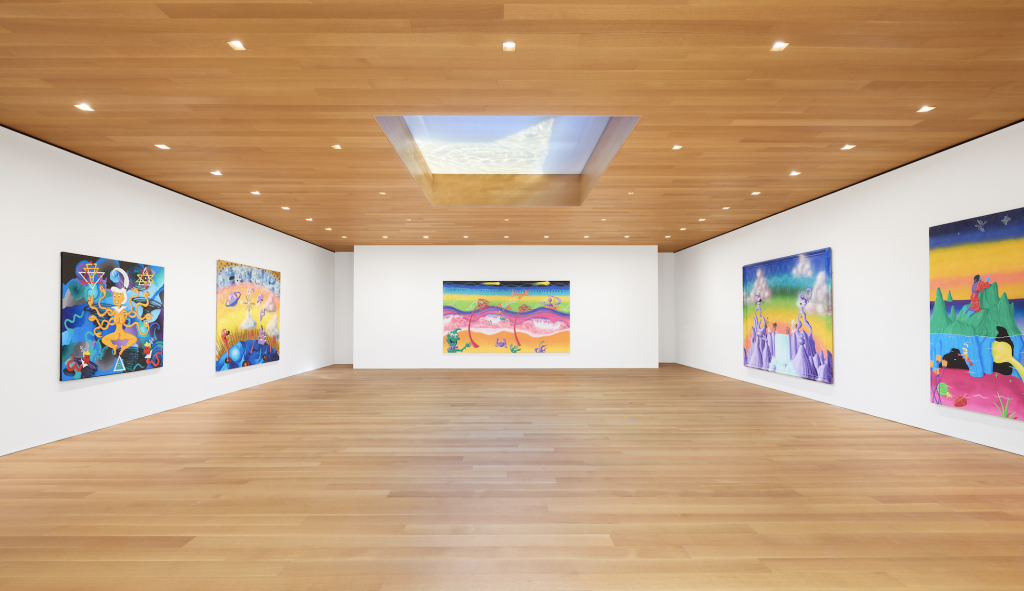 An installation photograph of Kenny Scharf at the Brant Foundation, featuring three white walls each hung with their own colorful painting, beneath a wooden ceiling with a square skylight.