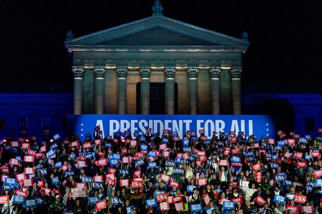 People in the Audience wait for Democratic presidential nominee, U.S. Vice President Kamala Harris, to speak during the closing rally of her campaign at the base of the iconic "Rocky Steps" at the Philadelphia Museum of Art on November 05, 2024 in Philadelphia, Pennsylvania. It is night and they are in front of the neo-classical museum with its many columns and pediment. There is a large blue sign with white lettering reading "a president for all" behind the crowd. 