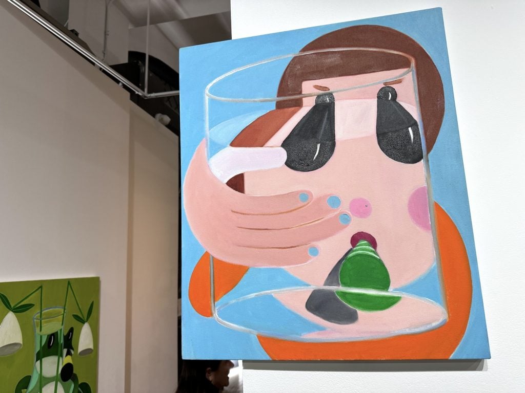 A colorful painting depicting an abstract human face with exaggerated features. The person appears to be holding a glass with water and a green object inside, which distorts parts of their face. The background is blue, and the figure is wearing an orange garment. A second artwork with a green theme is partially visible in the background.