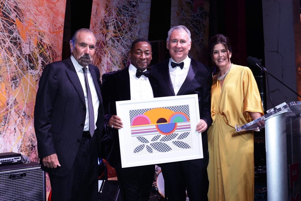 A photograph of a group of four people in formal attire all smiling and holding a colorful print of an artwork.