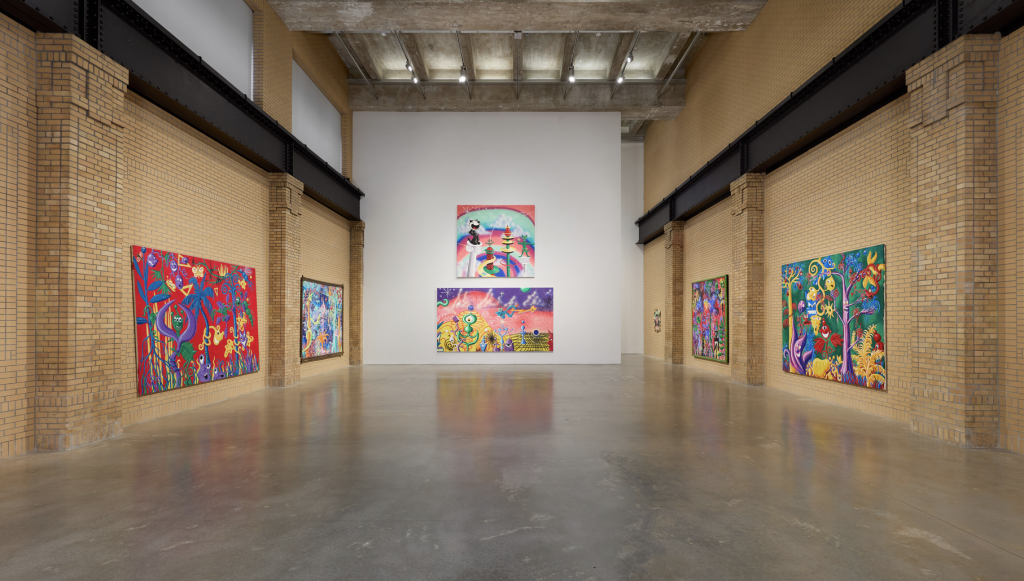 An installation photograph of Kenny Scharf at the Brant Foundation, featuring six colorful paintings hung amongst three walls, two of which are yellow brick while the back one is white.