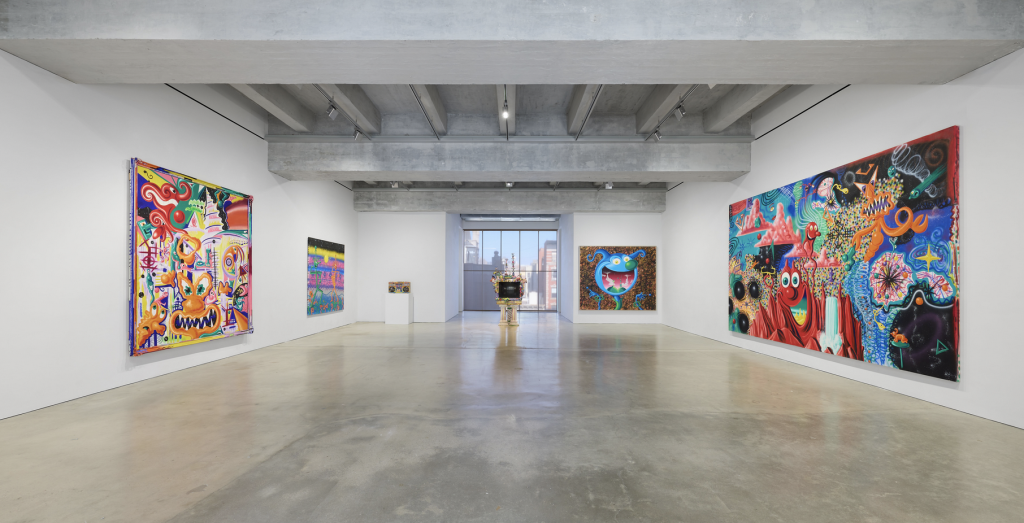 A photograph of an airy gallery displaying Kenny Scharf at the Brant Foundation, featuring colorful paintings on white walls and a freestanding TV sculpture before a window looking out on lower Manhattan.