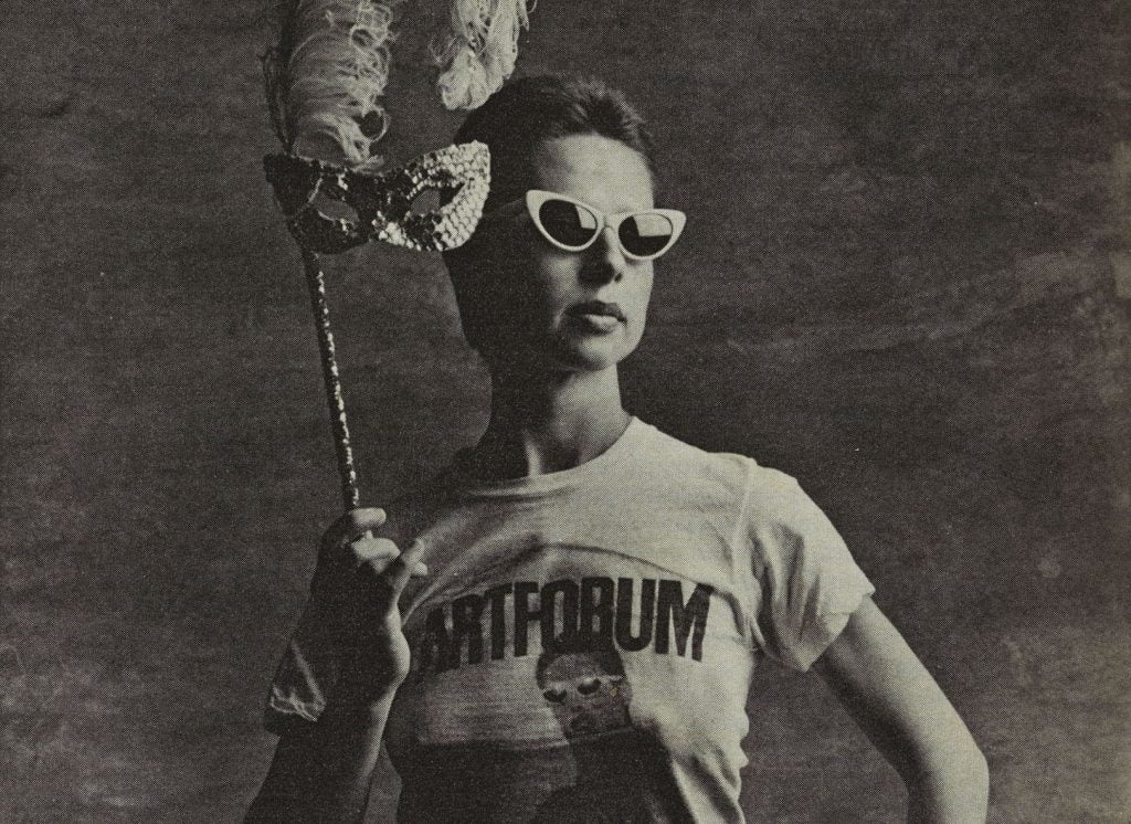 A black and white photo of Lynda Benglis wearing sunglasses and a t-shirt featuring her Artforum ad, while holding a mask.