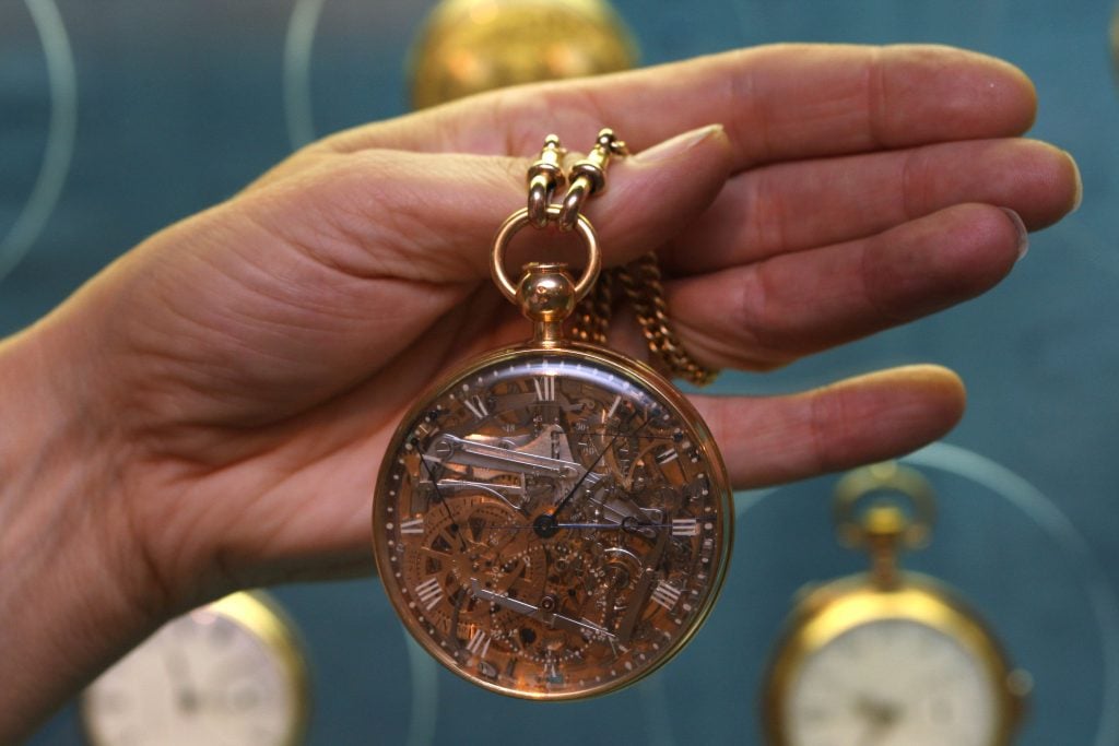 an intricately designed golden watch with a crystal dial and decoration made from precious stones held by a human hand against a blue background with other watches