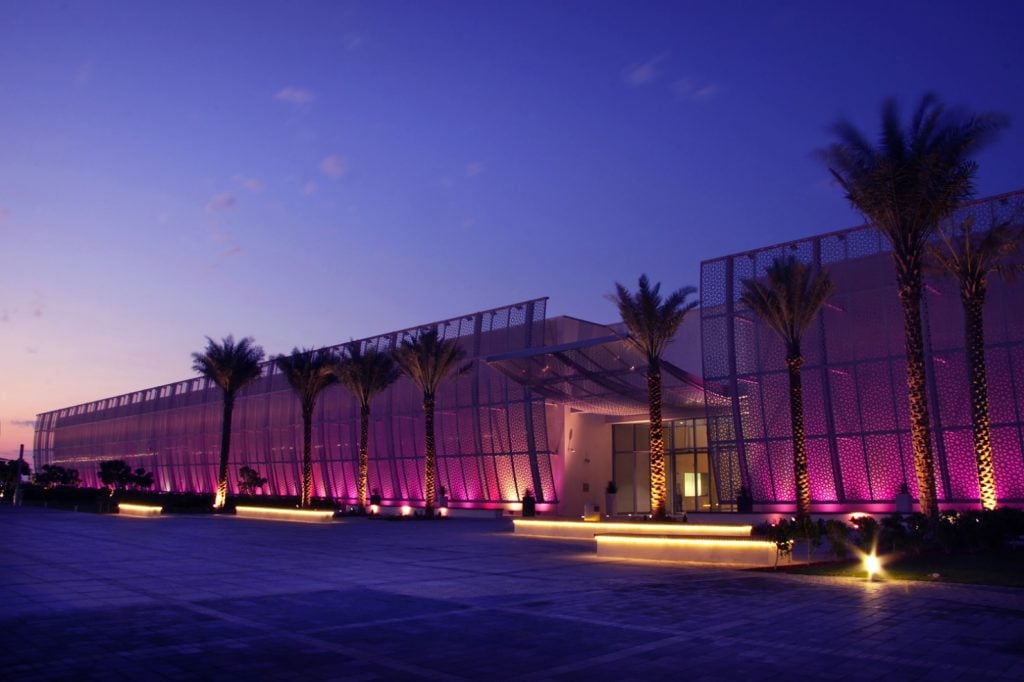 Exterior view of the convention center hosting Abu Dhabi Art lit up at night by purple lights and with palm trees lining the front.