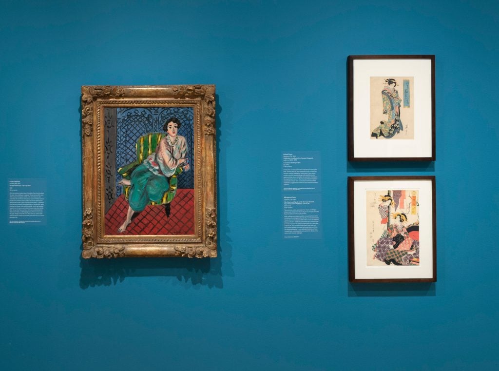 framed painting of a woman in colorful clothing on a blue museum wall next to two smaller framed woodblock prints of women with small texts in white ink