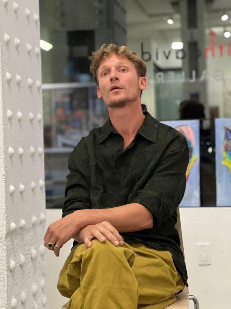 Portrait of artist Matthijs Scholten sitting down with his arms overlain wearing a black button down and khakis in front of a darkened window.