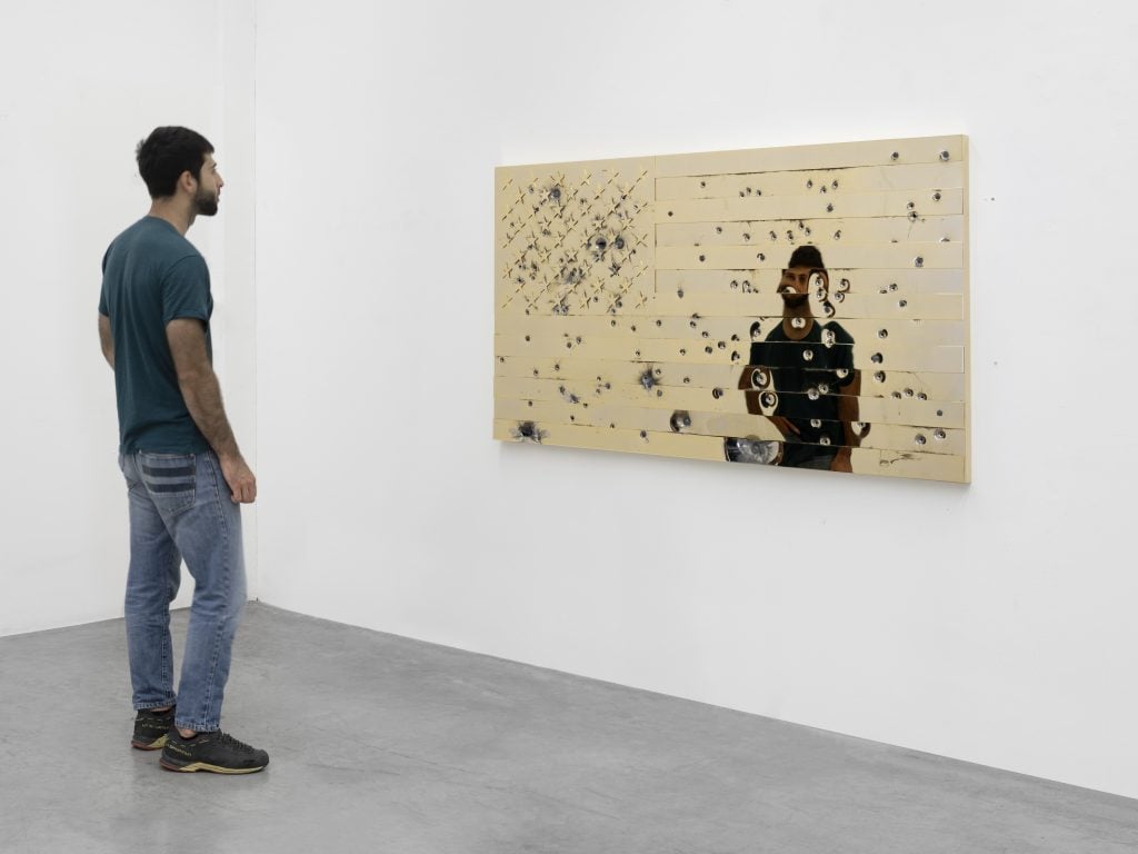 A man stands in front of a gold-plated panel with a US flag design that has been shot with bullets