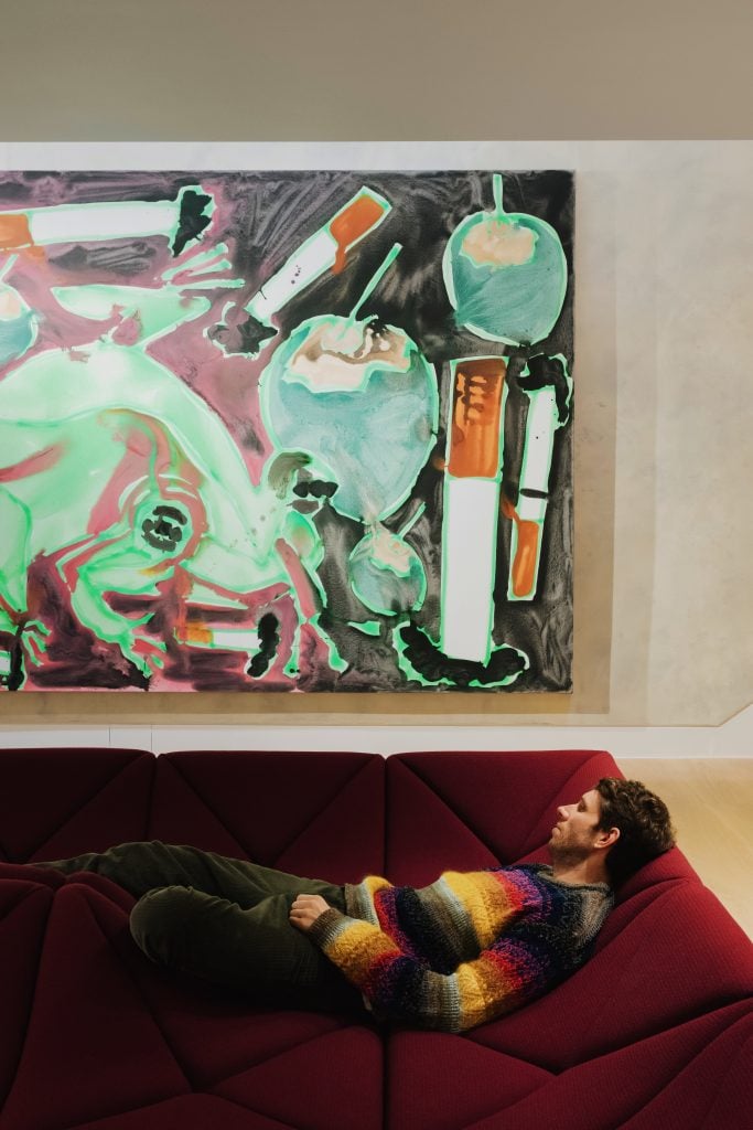 Dark Green founder Michael Doohan laying on a red velvet sofa underneath a painting by Katherine Bernhardt.