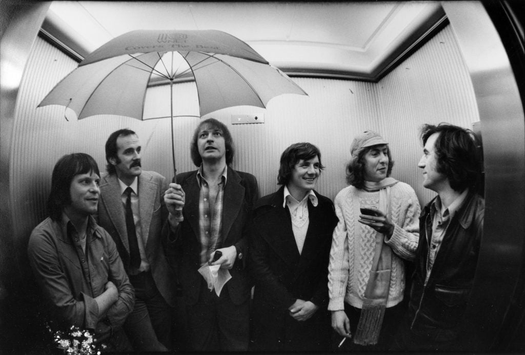 a black and white photograph of six men stading in an elevator, one has an umbrella open