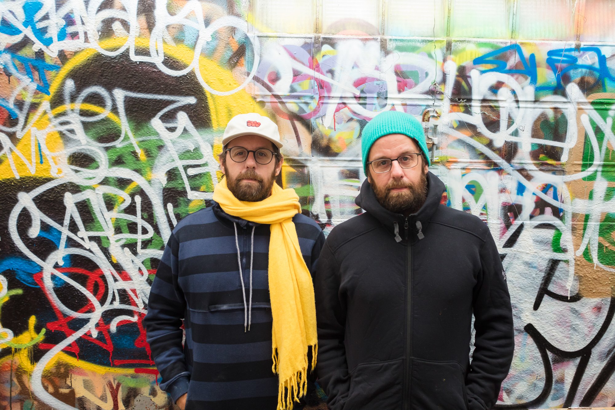 two men in black jackets wearing a yellow scarf and a bright blue hat standing in front of a wall filled to the brim with several layers of colorful graffiti