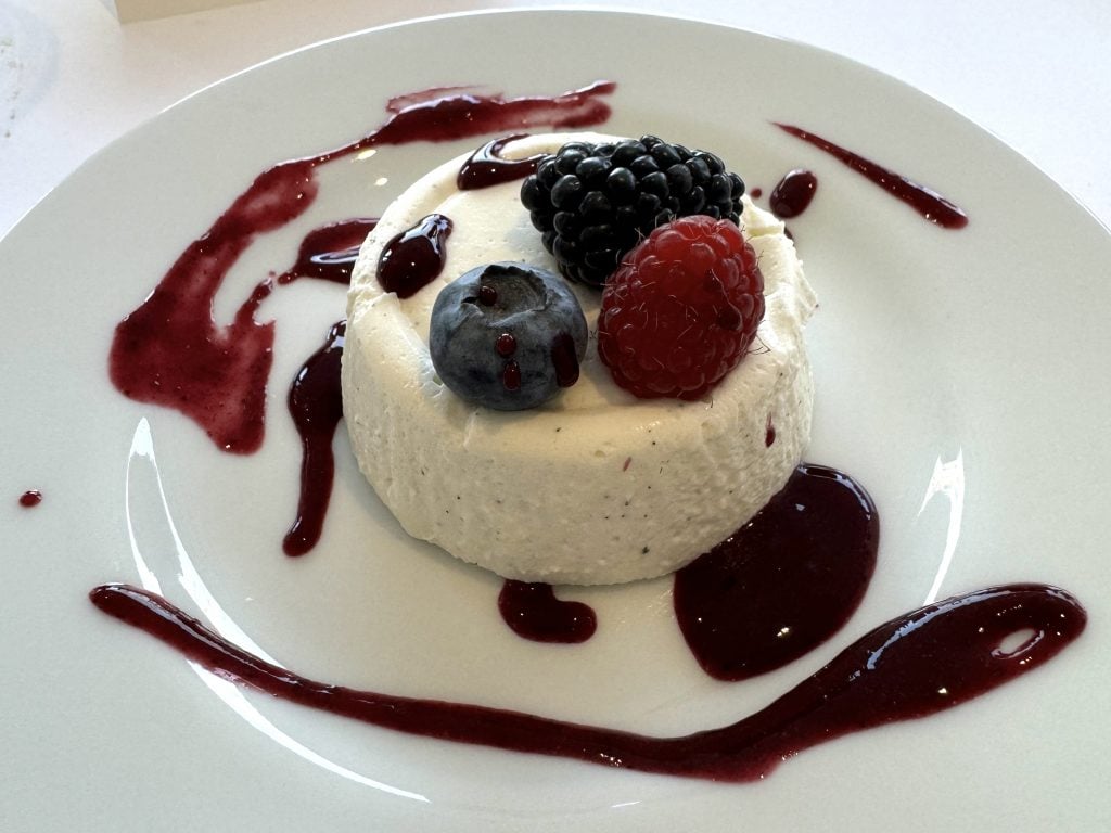 The panna cotta with berries by chef Luca Galli at Café Silvia at Magazzino Italian Art in Cold Spring, New York. It is a round white molded pudding in the center of a white plate, topped with a large blueberry, blackberry, and raspberry, and drizzled with a dark red berry sauce. 