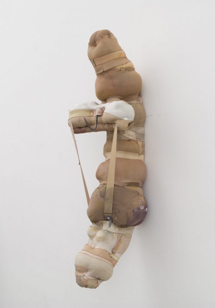 A wall-mounted sculptural assemblage by Patricia Ayres of brown and beige fabric stuffed with straps holding it together.