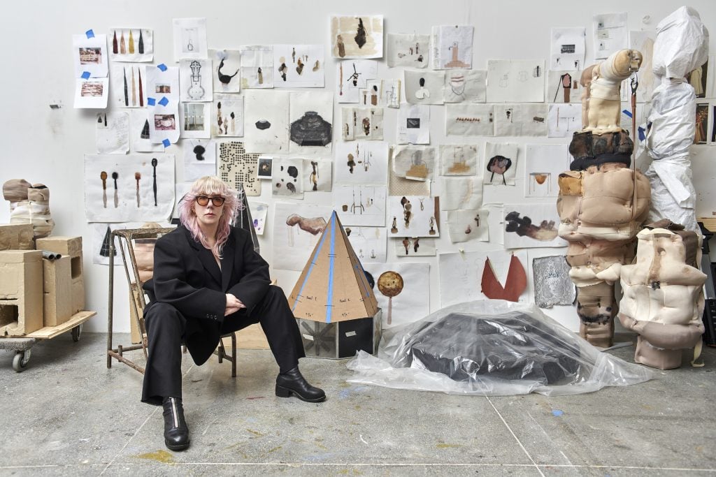 American artist Patricia Ayres in a black oversized suit sittin in a gallery space with the back wall covered in small-scale drawings and watercolors.