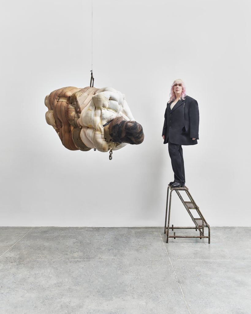 American artist Patricia Ayres wearing an all black oversized suit standing on a three-step step ladder next to one of her suspended assemblage sculptures against a white wall and light concrete floor.