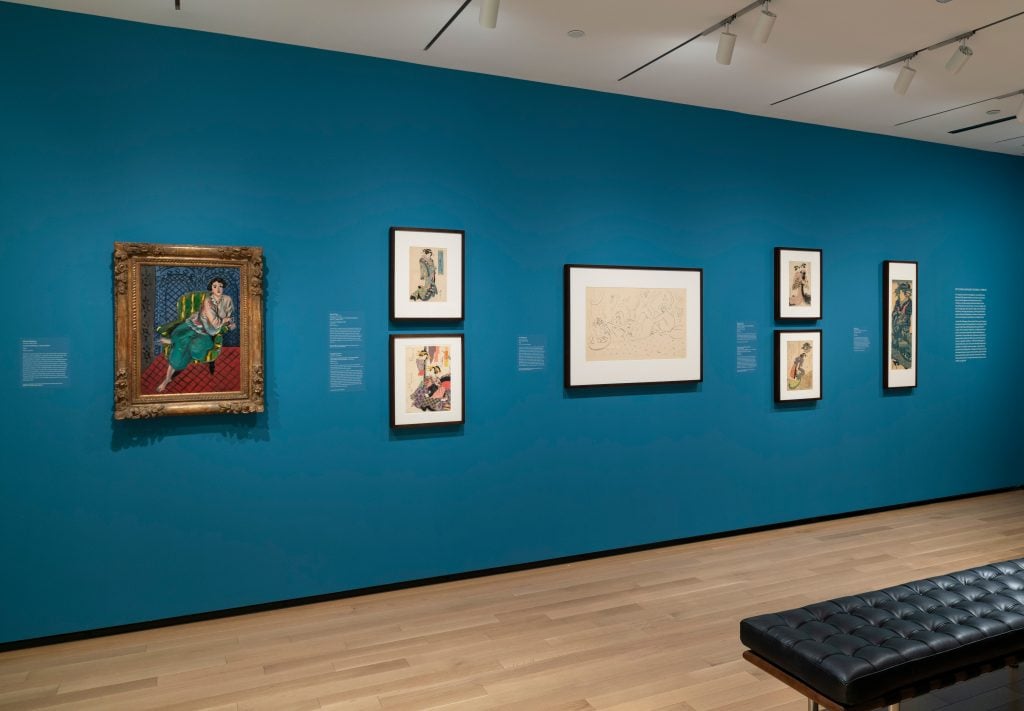 a collection of paintings and woodblock prints in fancy frames attached to a blue wall inside a museum exhibition hall without any people around