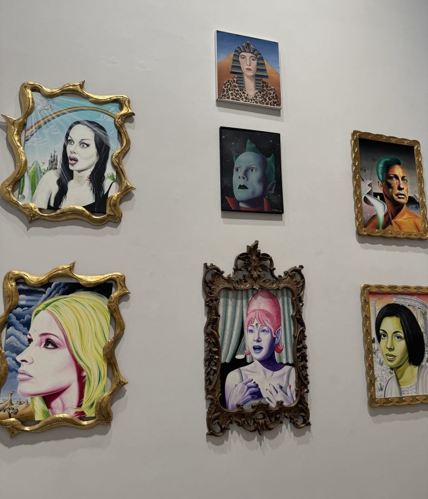 A photograph depicting a grid of seven small framed painted portraits arranged on a white wall.