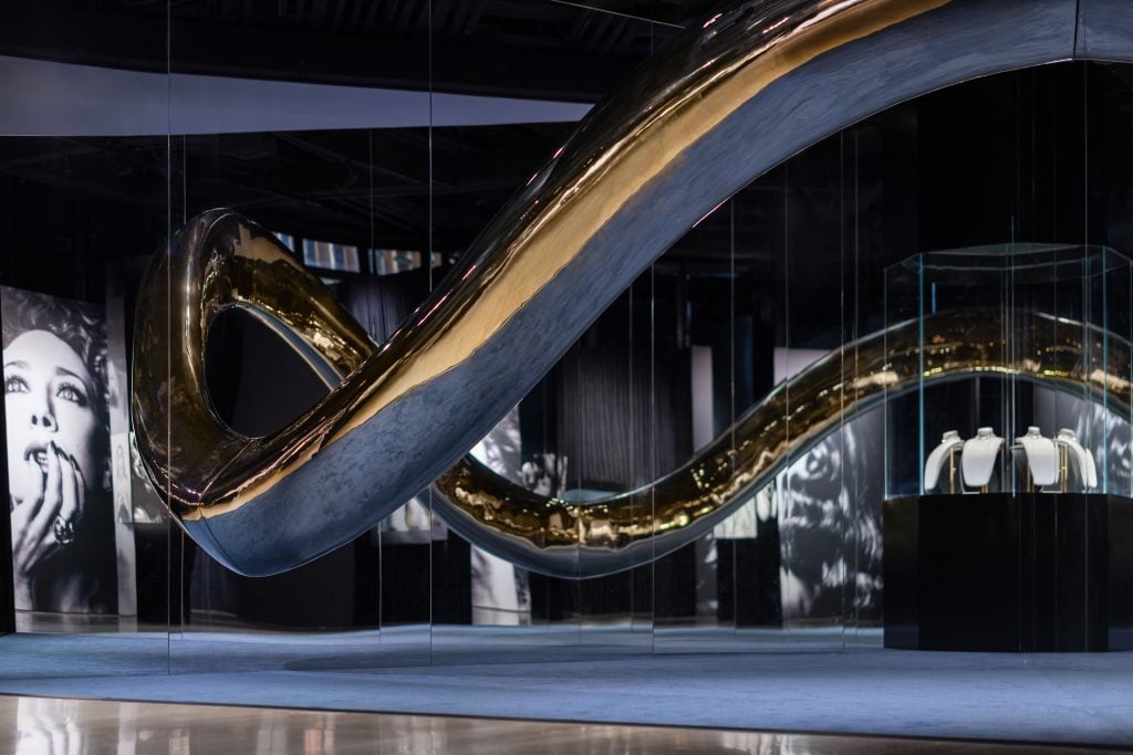 A photograph of a dark, dramatic room featuring black and white photos as well as a large silver rendition of the Pomellato wave necklace inside the brand's retrospective.