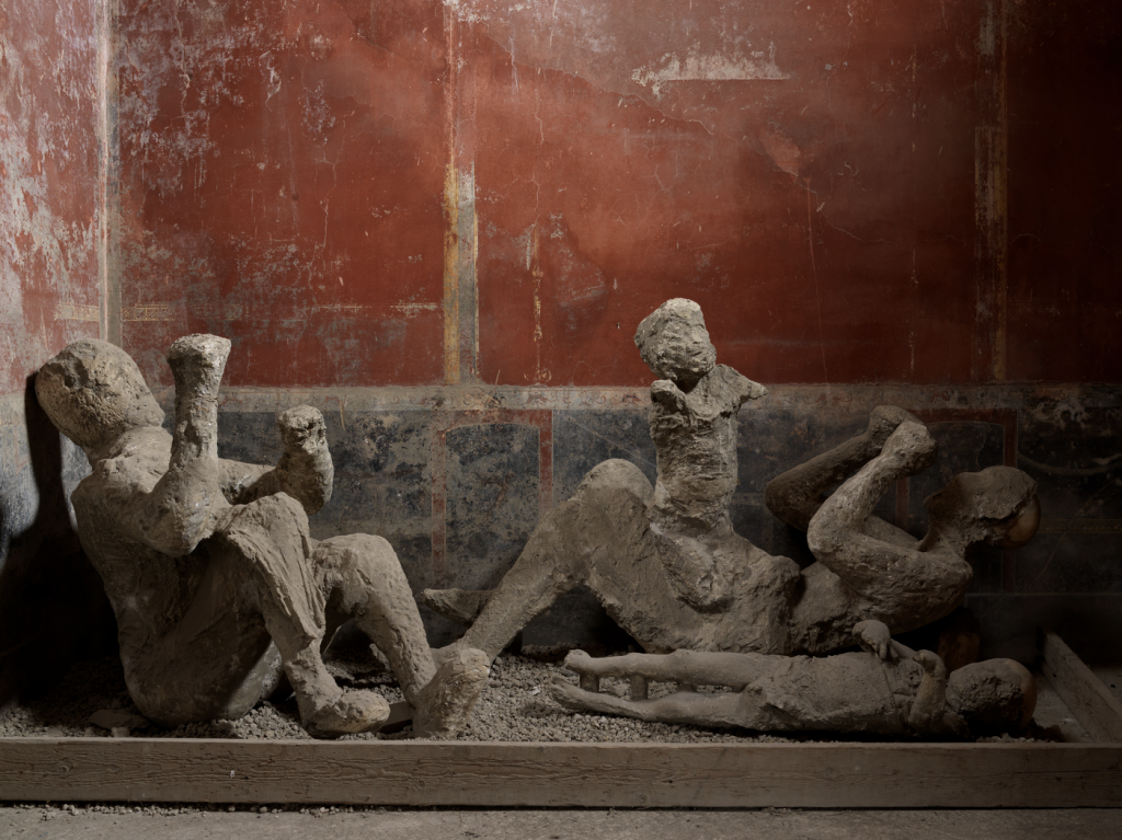 A photograrph of a group of plaster casts of Pompeii victims situated throughout a red-tinged room.