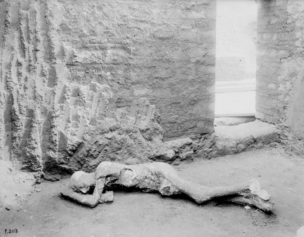 A black and white photo of the cast of a Pompeii victim lying on the ground before the corner of a brick building