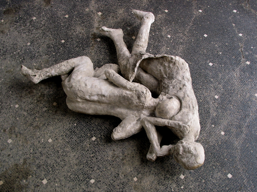 A photograph taken from above, looking down on the casts of two Pompeii victims locked in an embrace atop a grey floor.