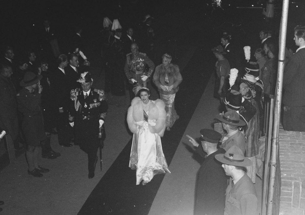 Princess Margaret in a white gown and shiny tiara walks down a carpet flanked by a military man