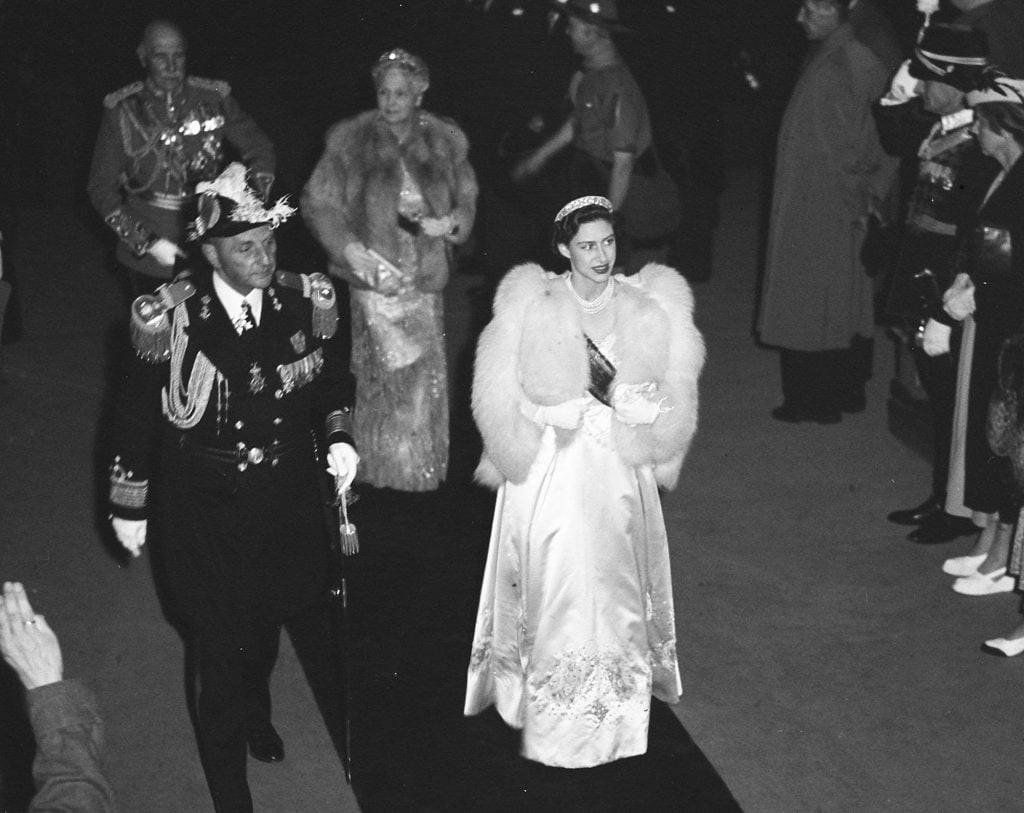 Princess Margaret in a white gown and shiny tiara walks down a carpet flanked by a military man