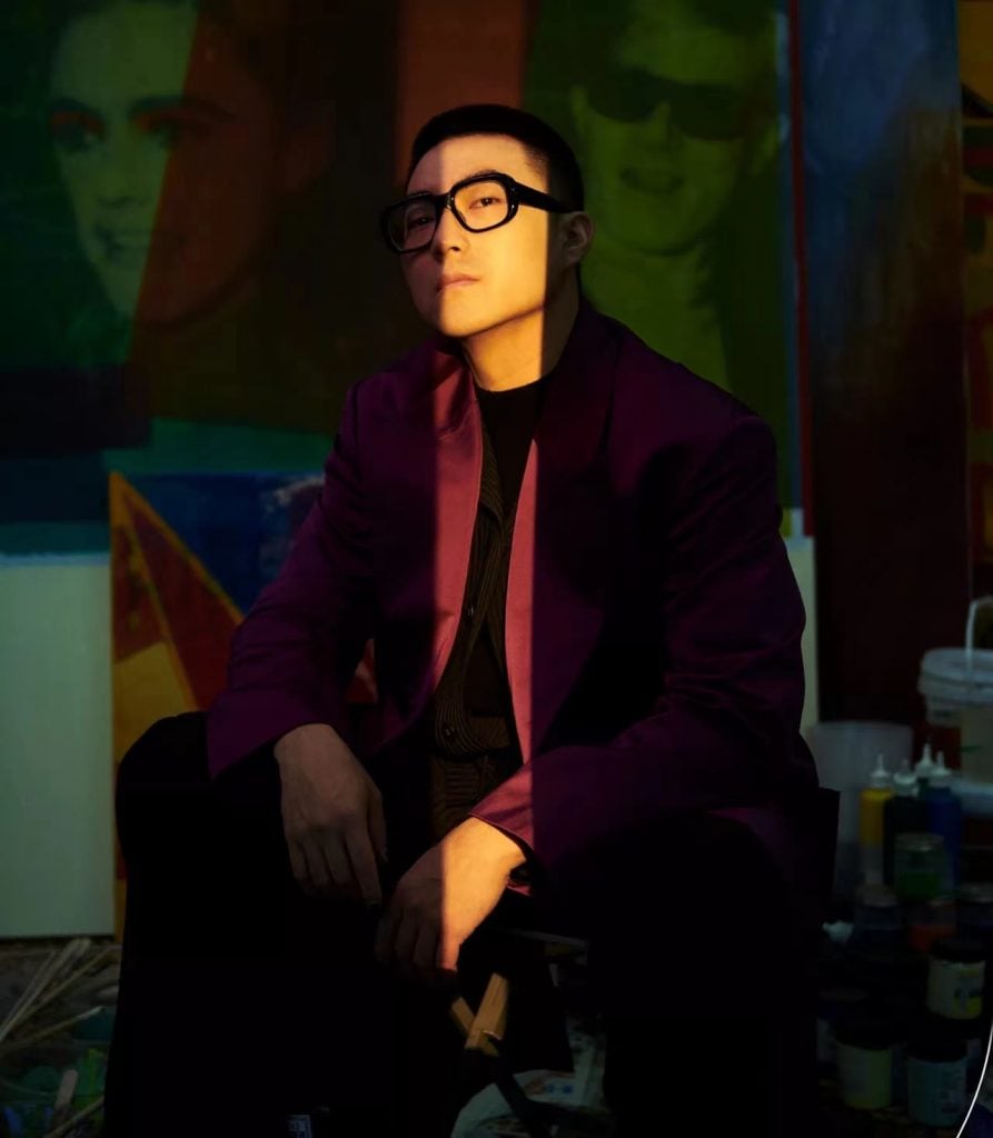 Portrait of a person seated in a creative studio, wearing a purple blazer and glasses, illuminated by dramatic lighting with vibrant artwork in the background.