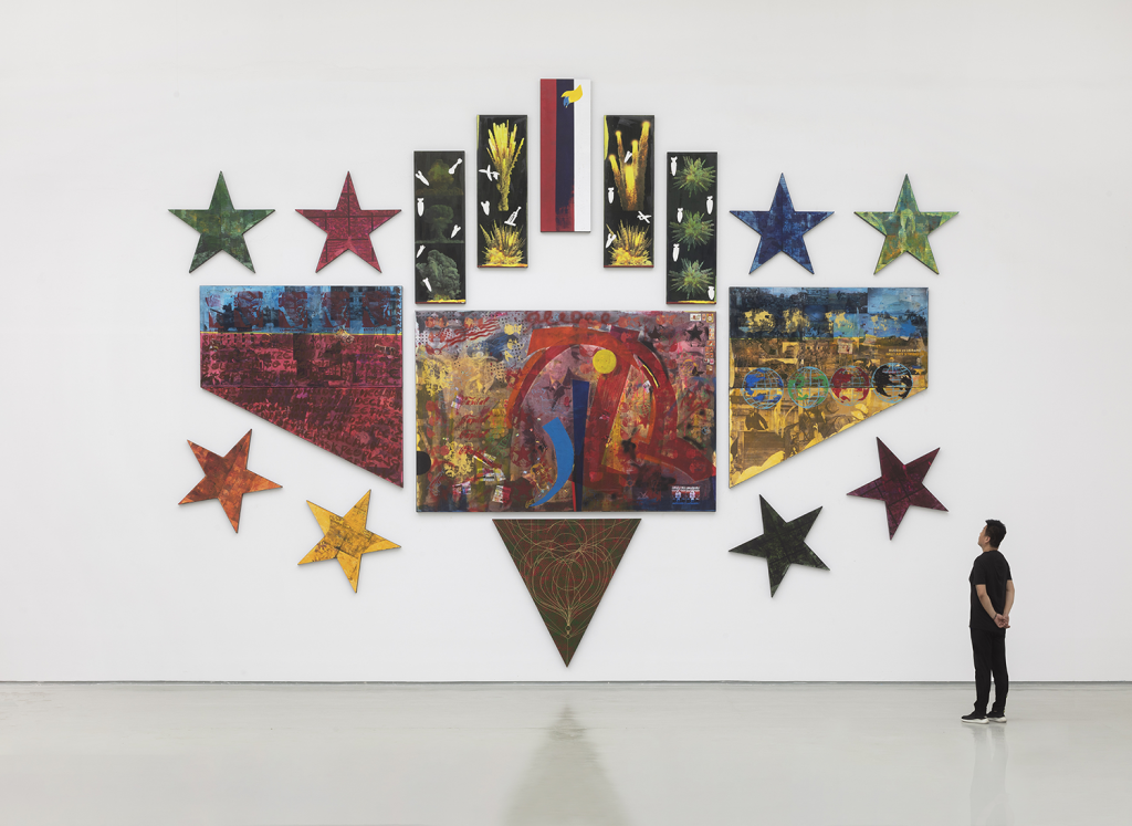 Large multi-panel artwork featuring vibrant abstract paintings, colorful star shapes, and geometric patterns, observed by a person standing in a spacious white gallery.