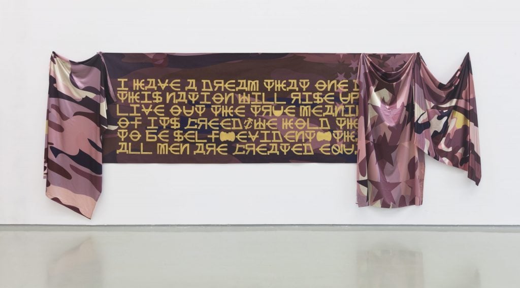 Textile artwork featuring draped fabric in camouflage patterns, with bold golden text quoting a historical speech, displayed on a white wall in a gallery.