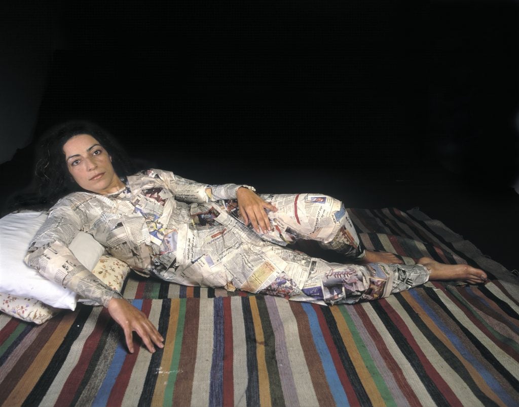 Self-portrait of Raeda Saadeh lounging on a striped blanket on a white pillow against a background of black, fully enrobbed in cutouts from the pages of glossy magazines and newspapers.