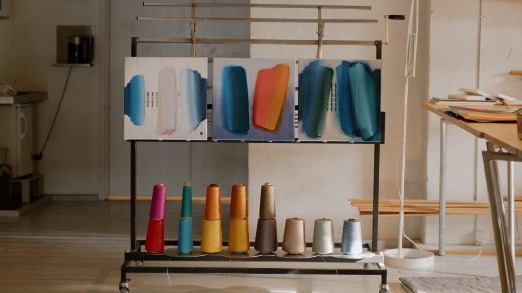 A photograph of four small square abstract artworks on stands above spools of colorful thread.