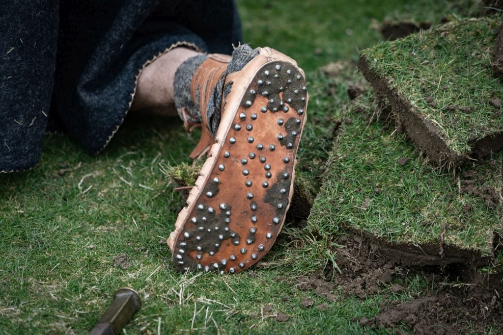 photograph of the underside of a shoe worn by someone laying down on grass, with the brown sole visible so the nails stuck through it show.